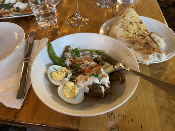 Bowl of authentic Israeli food with egg opened up in a white bowl and spoon. Pita is on the table.